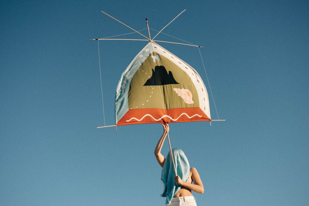 Person holding a large, house-shaped kite decorated with landscape art against a clear blue sky; their head is covered with a light blue cloth.