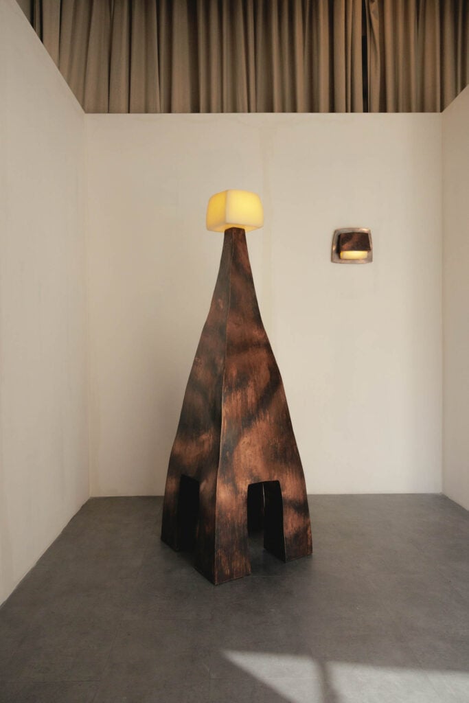 A tall, abstract wooden sculpture with a yellow light on top stands in a minimalist room with gray tile flooring and plain walls; a small wall light is mounted nearby.