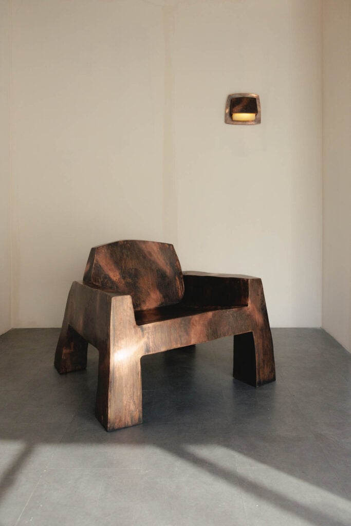 A sculptural, angular bronze chair sits in a minimalist room with a matching bronze wall sconce above and neutral-colored walls.
