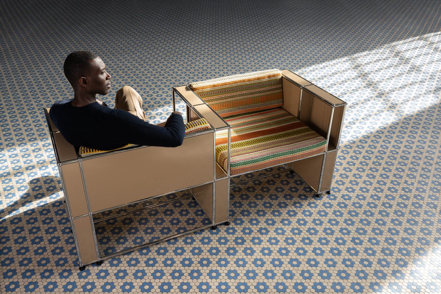 A man sits in a modern armchair next to an empty, striped armchair on a patterned tile floor with sunlight streaming in.
