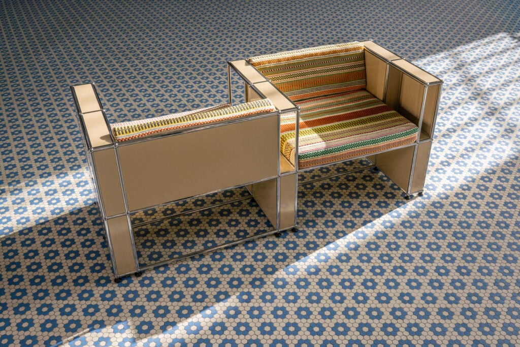 A modern chair with a metallic frame and colorful striped cushions sits on a blue and white geometric patterned tiled floor in sunlight.