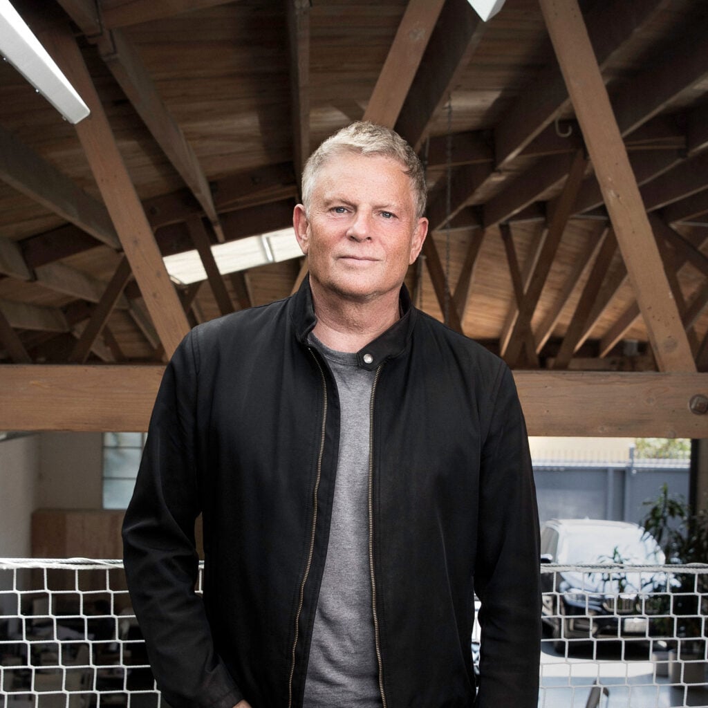 A man with short blonde hair wearing a black jacket and gray shirt stands indoors in front of wooden beams and a netted barrier.