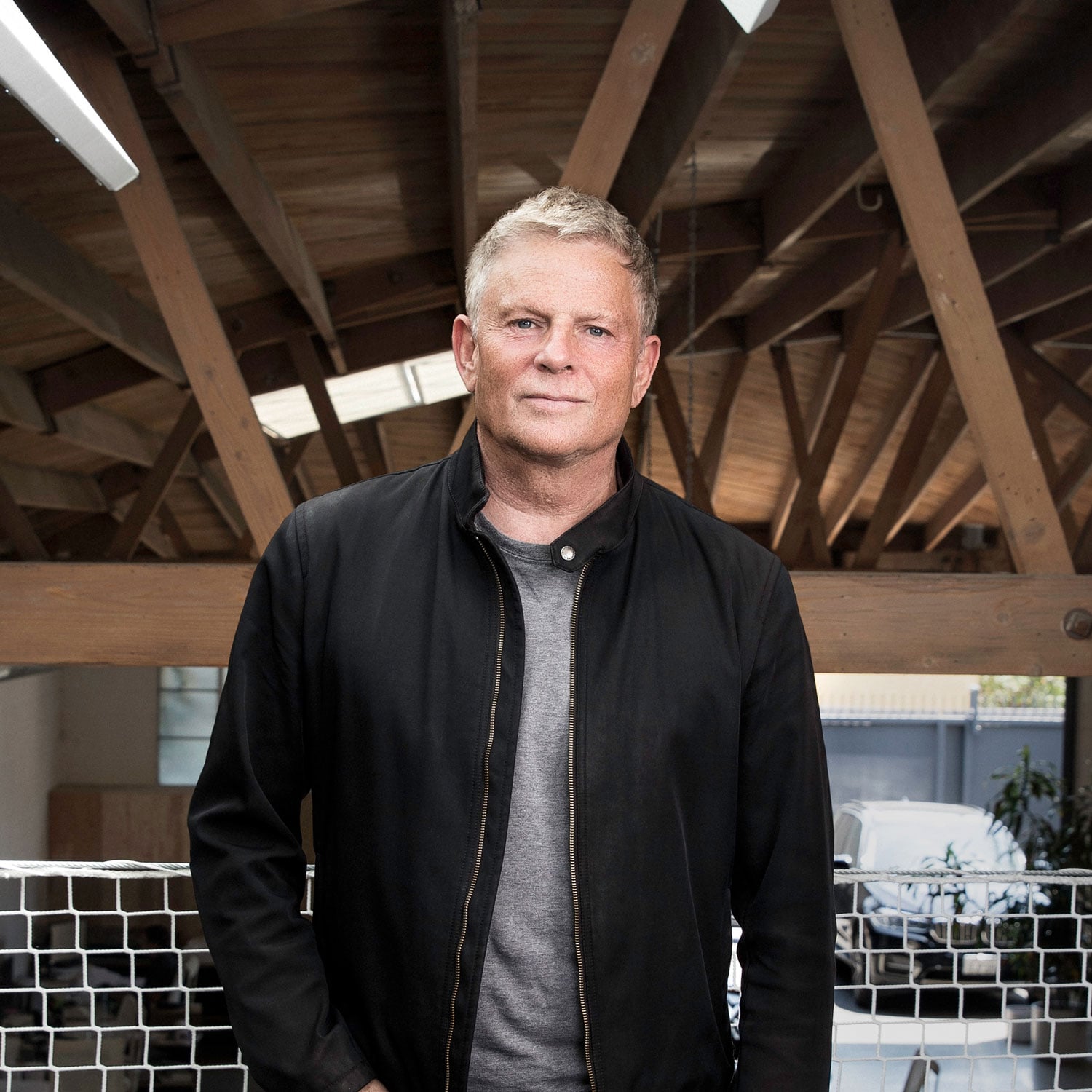 A man with short blonde hair wearing a black jacket and gray shirt stands indoors in front of wooden beams and a netted barrier.