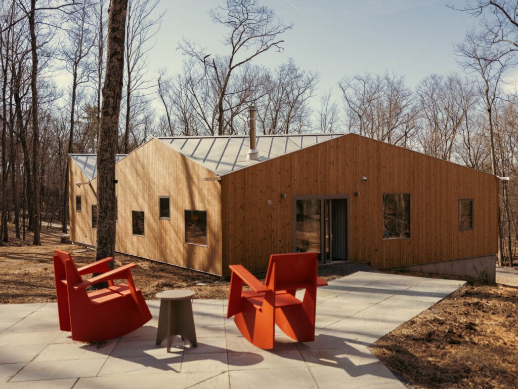 A modern, wood-clad house in a forest setting with two orange chairs and a gray table on a patio.