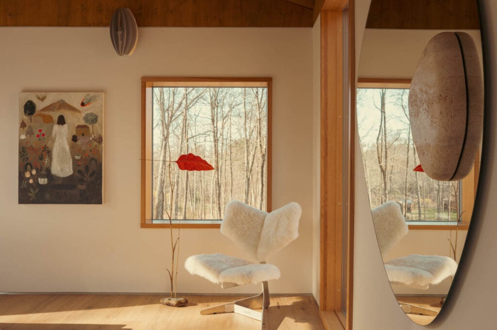 A modern room with a white fur chair, abstract painting, tall red lamp, large round mirror, and a window showing bare trees outside. Warm sunlight fills the space.