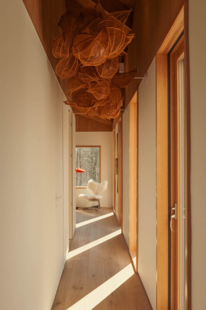 A narrow hallway with wood floors and white walls, featuring sculptural leaf-like art on the ceiling, leading to a bright room with a white chair by large windows.