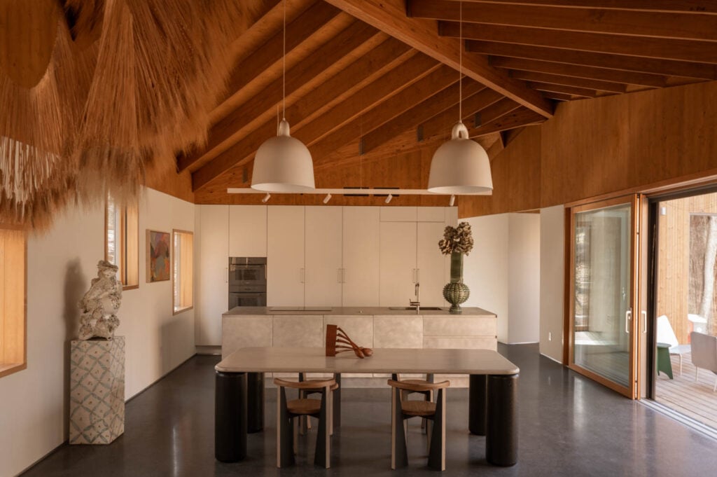 Open-plan kitchen with exposed wooden ceiling, white cabinetry, central island, two pendant lights, modern art, and sliding glass doors leading to an outdoor seating area.