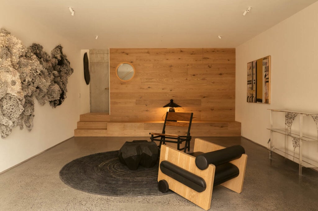 Minimalist room with a wooden accent wall, modern black chair, round rug, abstract wall art, a round mirror, lamp, and a geometric black table on a polished concrete floor.