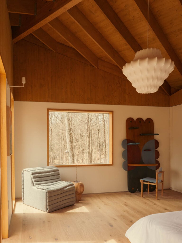 A modern bedroom with wood ceiling and floors, a large window overlooking trees, a gray lounge chair, a unique wall-mounted mirror, and a white pendant light.
