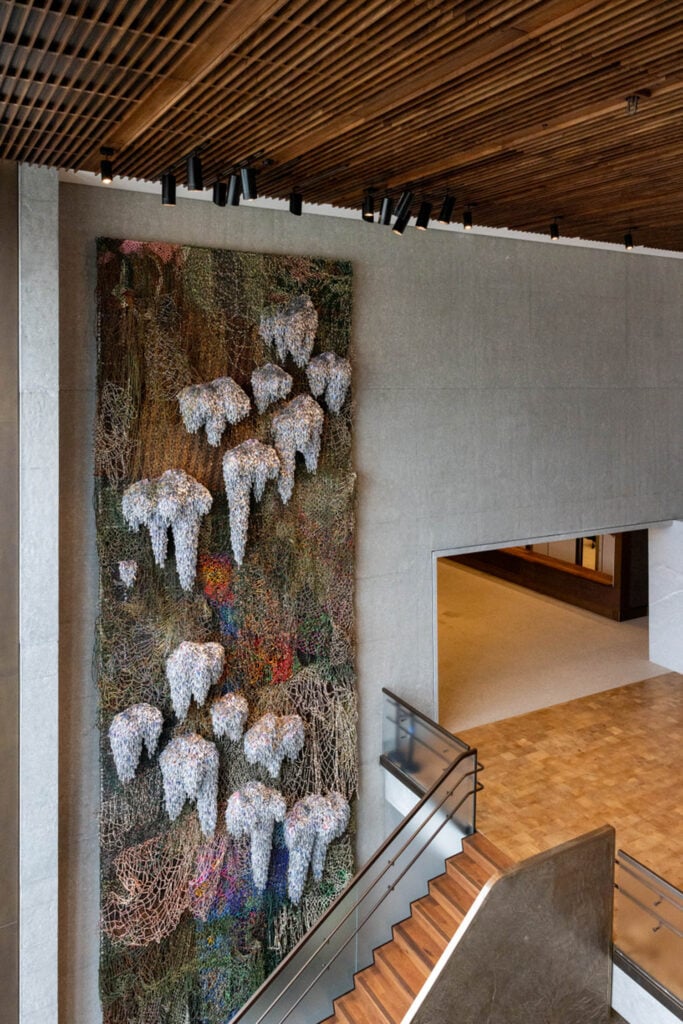 Large vertical mixed-media wall sculpture with organic shapes and clustered white forms displayed in a modern building interior near a staircase and wood-paneled ceiling.