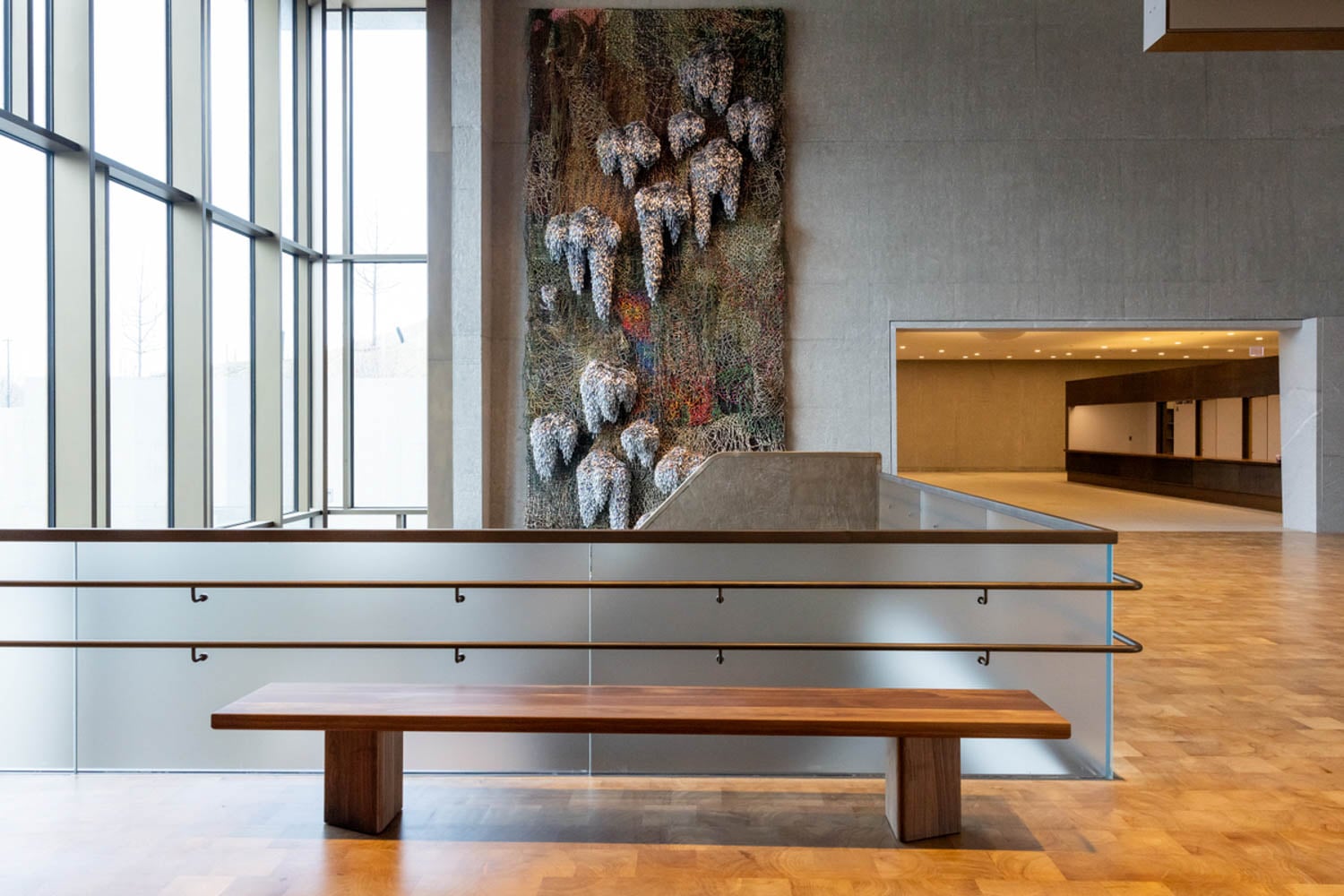 A modern lobby with large windows, a wooden bench, glass railing, and a textured wall art piece featuring abstract organic shapes.