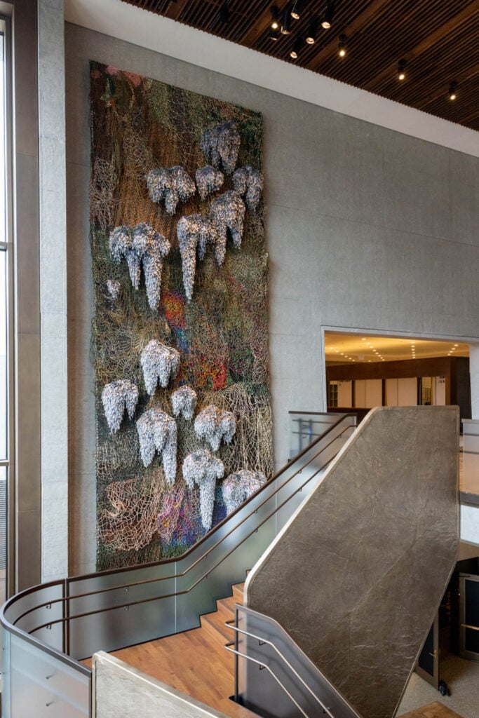 Large abstract wall sculpture featuring clusters of metallic and netted materials installed beside a staircase in a modern building lobby.