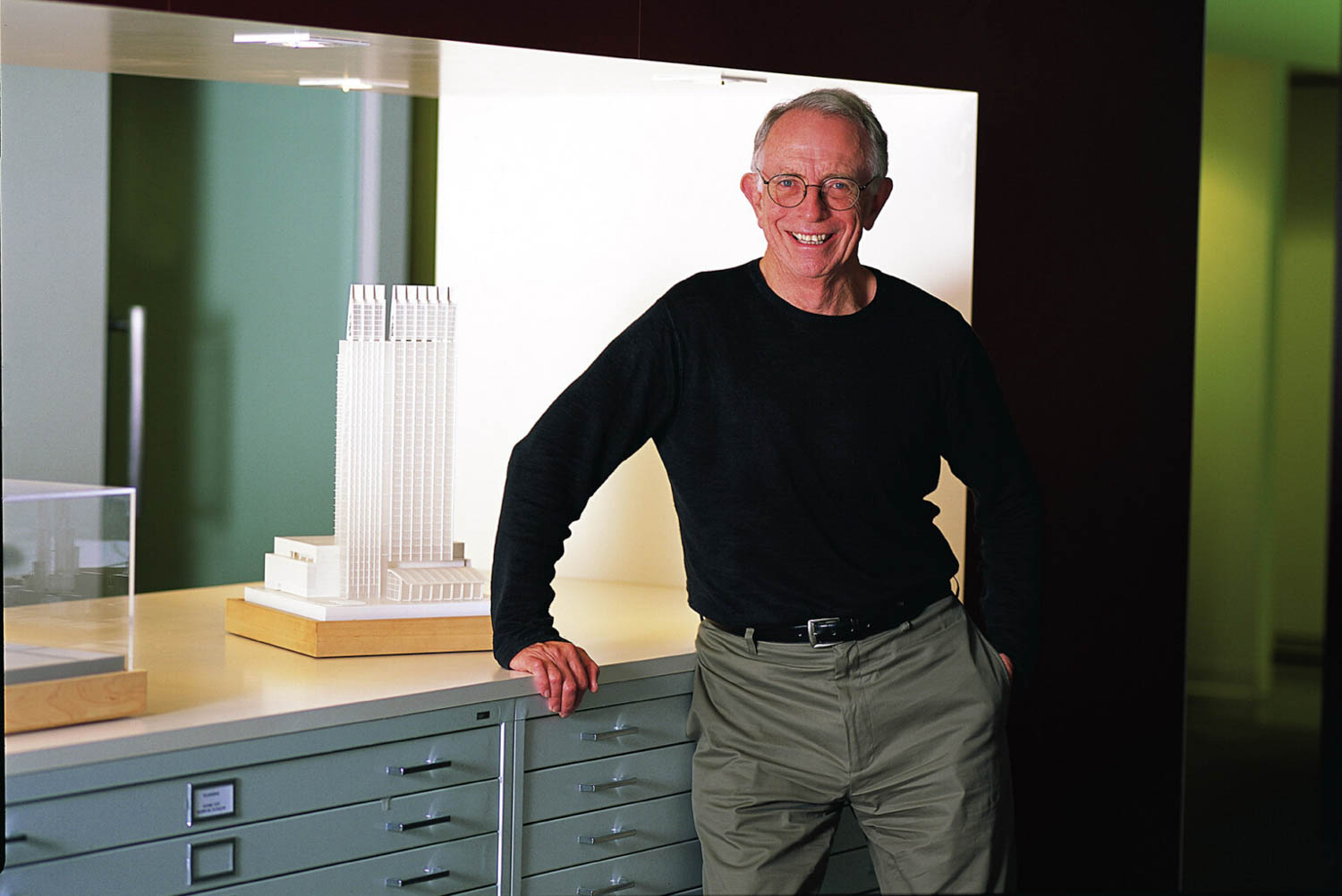 An older man wearing glasses and a black shirt stands and smiles beside a scale architectural model of a tall building on a cabinet.