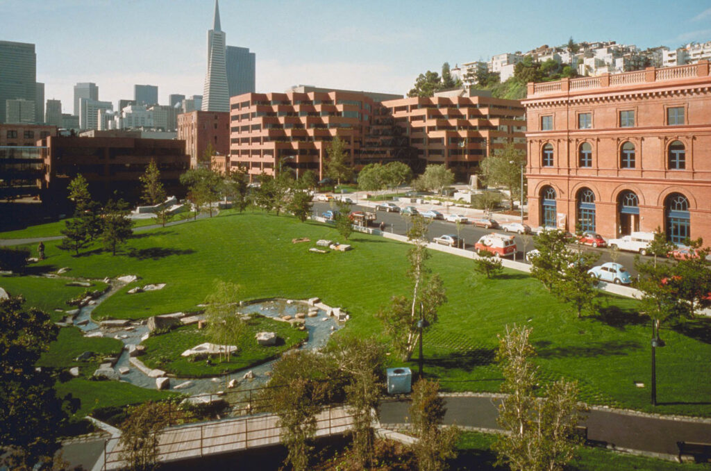 A city park with a stream and green lawn is surrounded by red brick buildings and parked cars, with a city skyline in the background.