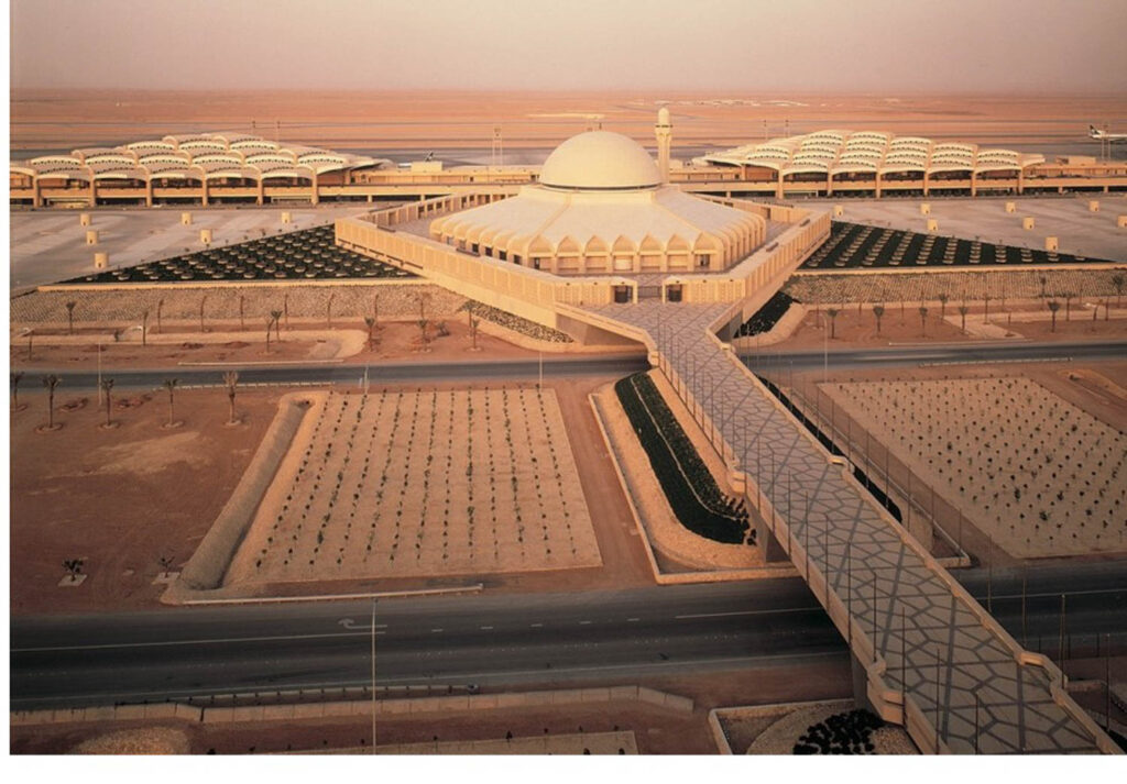 An aerial view of a large, modern airport terminal designed by Bill Valentine reveals a central domed building, extensive landscaping, and several covered structures in a striking desert setting.