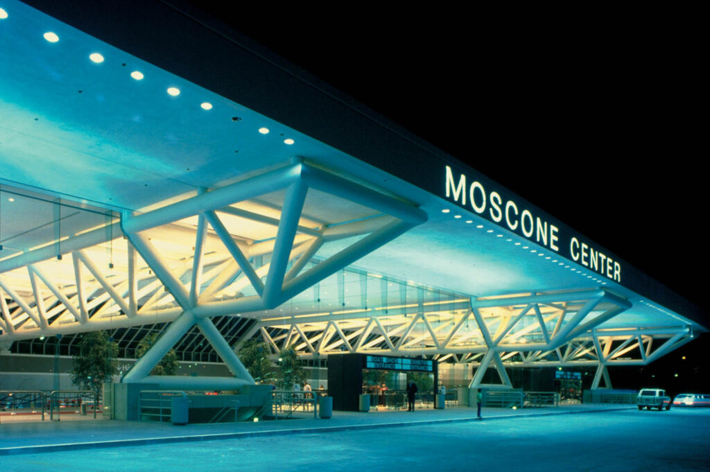 Night view of the illuminated Moscone Center entrance, featuring modern architectural beams inspired by Bill Valentine, glass walls, and visible signage above the entryway.