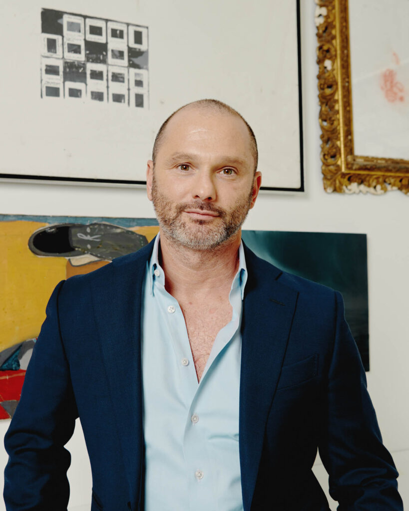A man in a navy blazer and light blue shirt stands in front of a wall with various framed artworks.