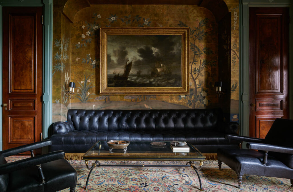A dark leather sofa and armchairs by Billy Cotton sit in a classic room with wood-paneled doors, patterned wallpaper, a glass coffee table, and a large framed painting above the sofa.