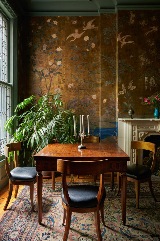 A wooden dining table with four chairs sits on a patterned rug beside a plant, with a decorative wall featuring birds and flowers, and a white fireplace with a vase of flowers—a cozy space inspired by Billy Cotton’s timeless designs.