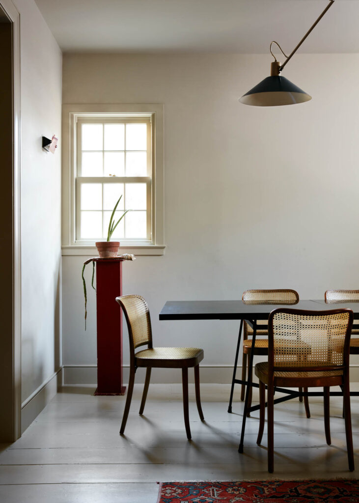 Minimalist dining room with a black table, cane chairs, and a potted plant on a red pedestal. A modern Billy Cotton lamp is mounted above, while natural light streams in through the window.