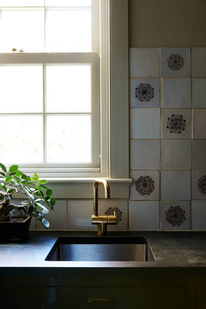 A kitchen sink with a gold faucet by Billy Cotton is set under a window. Floral patterned tiles cover the backsplash, and a potted plant sits on the counter.