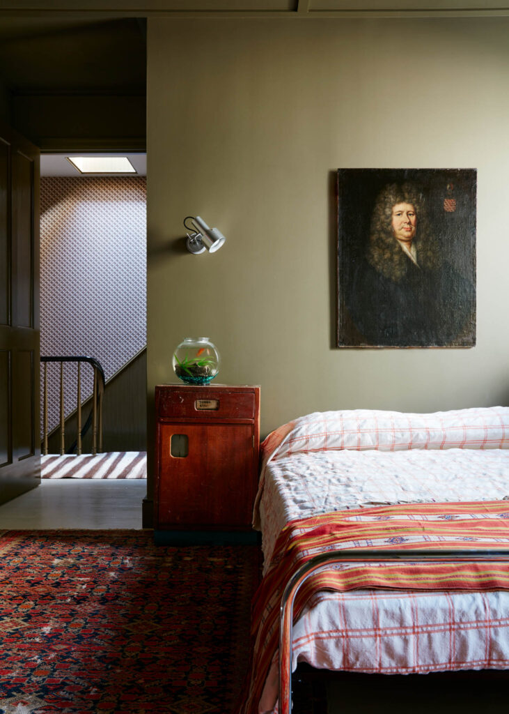 A bedroom with a striped bedspread, a red rug, and a wooden nightstand with a fishbowl showcases Billy Cotton’s signature style; an olive-green wall holds a portrait painting, and the hallway is visible in the background.