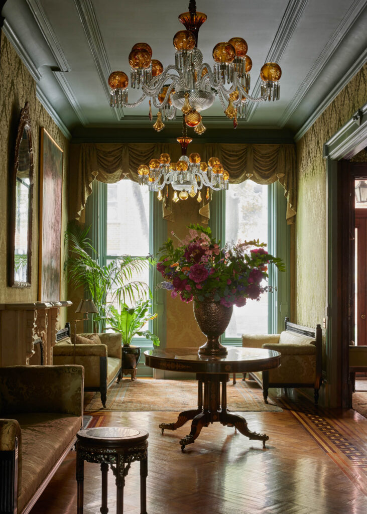 Ornate living room with chandeliers, tall windows with drapes, vintage furniture styled by Billy Cotton, and a round table with a large floral arrangement in the center.