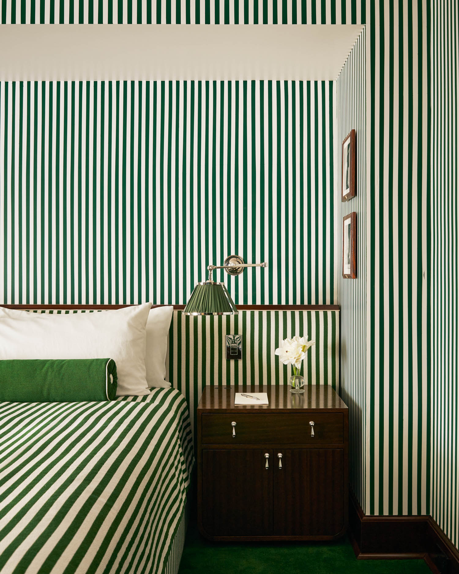 Bedroom with green and white vertical striped wallpaper and bedding, wooden nightstand with lamp, small vase of flowers, and two framed pictures on the wall.