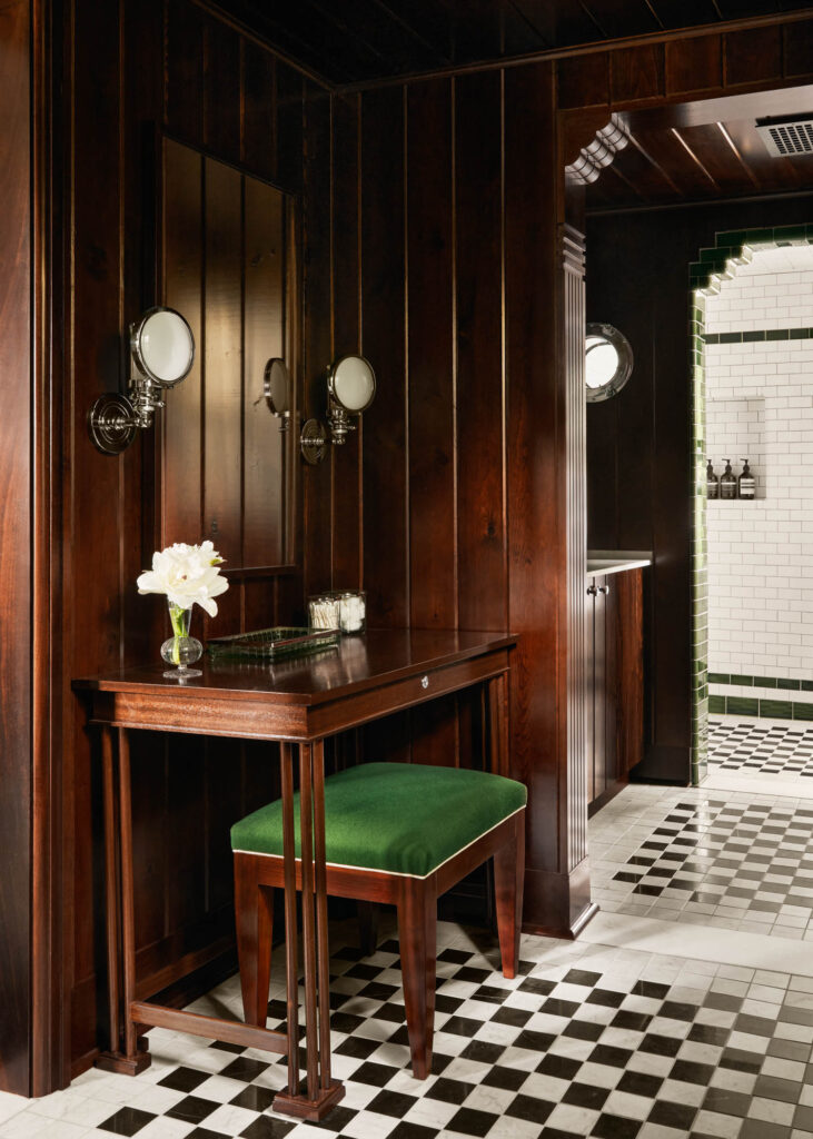 A wooden vanity with a green cushioned stool, wall-mounted mirrors, and a vase of white flowers, set in a wood-paneled room with black and white checkered floor tiles.