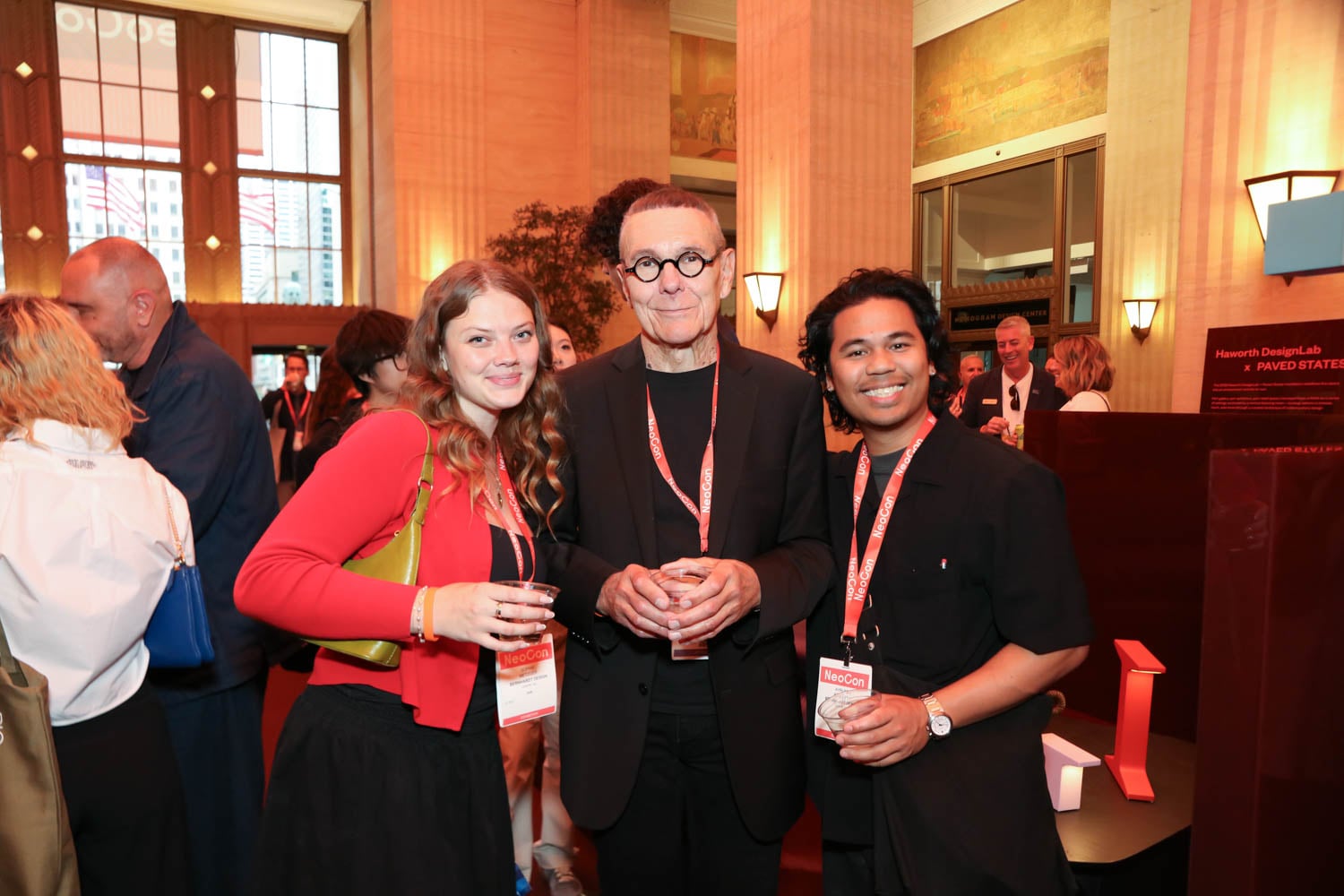 Three people wearing event badges pose together at an indoor reception, holding drinks, with other attendees visible in the background.