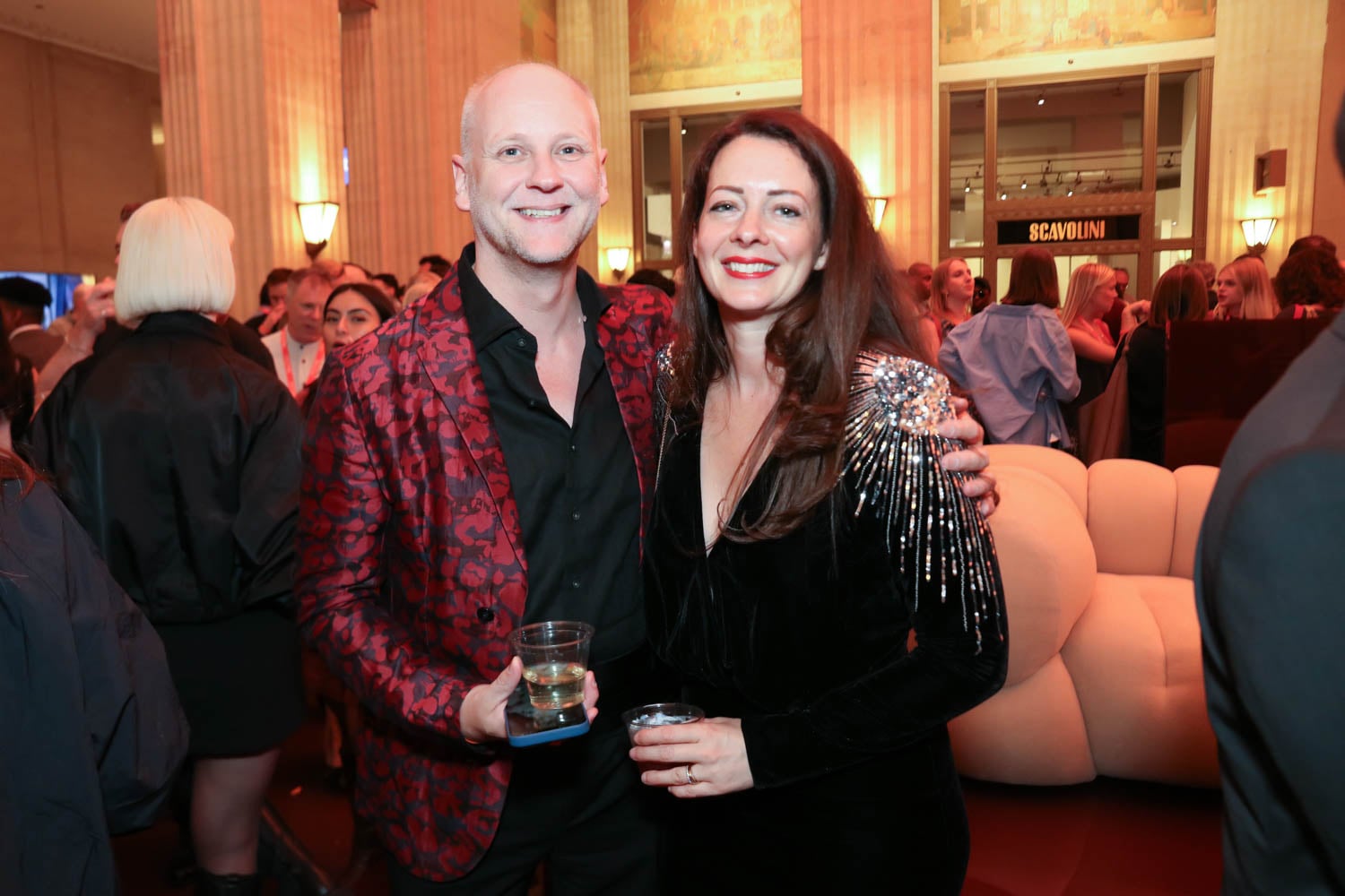 Two people stand closely together at an indoor event, both smiling at the camera; the man holds a drink, and the room is filled with other attendees in the background.