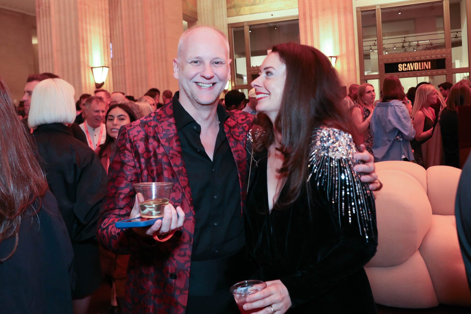 A man in a red patterned blazer and a woman in a black dress with sequin details stand together at a crowded indoor event, both holding drinks.