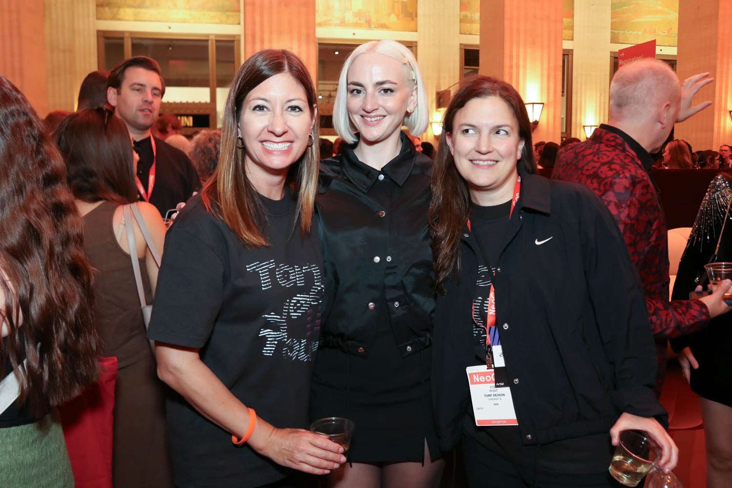 Three women pose together at an indoor event, wearing black outfits and name badges. Other people are visible in the background.