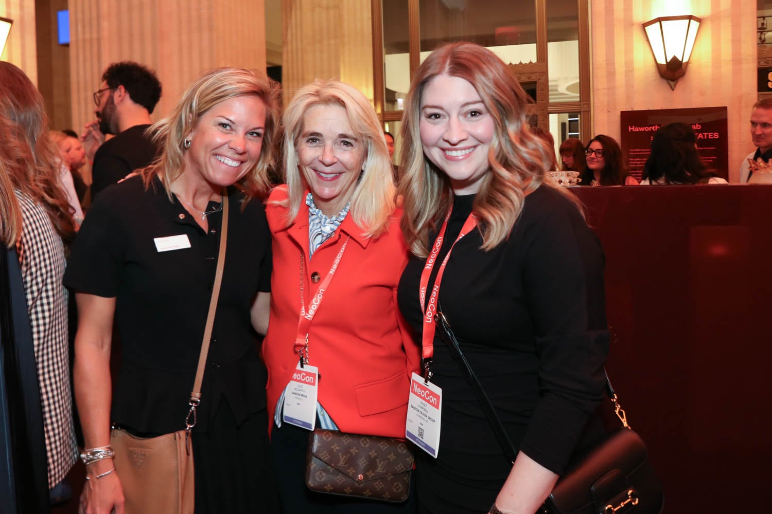 Three women stand together and smile at an indoor event, each wearing name badges and conference lanyards, with other attendees visible in the background.