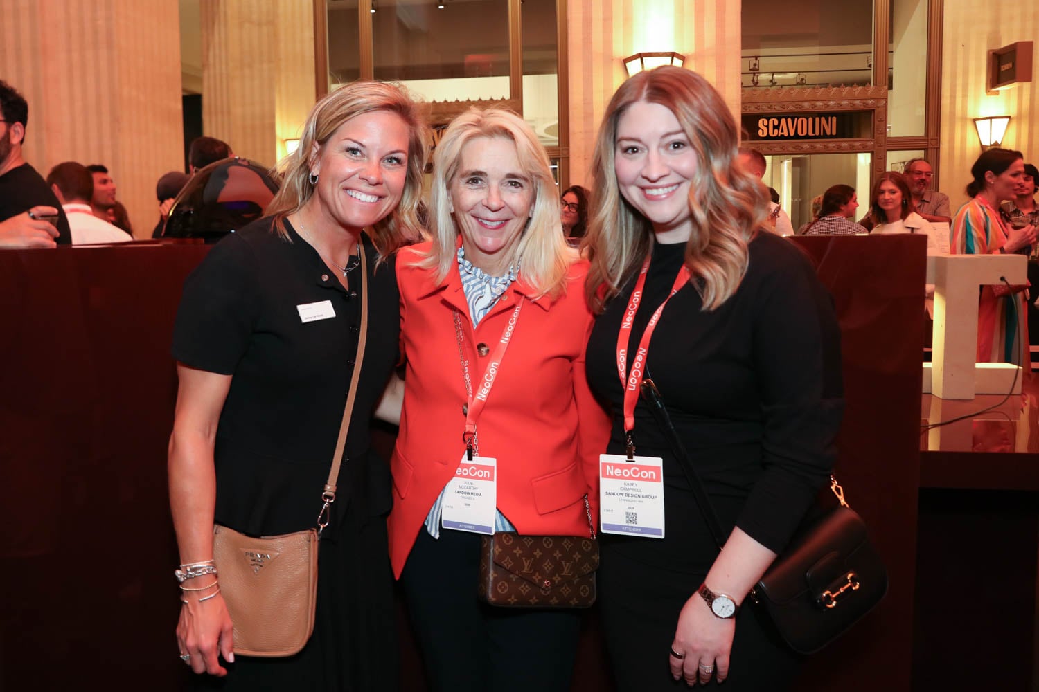 Three women stand together indoors at an event, smiling at the camera and wearing NeoCon badges around their necks. Other attendees are visible in the background.