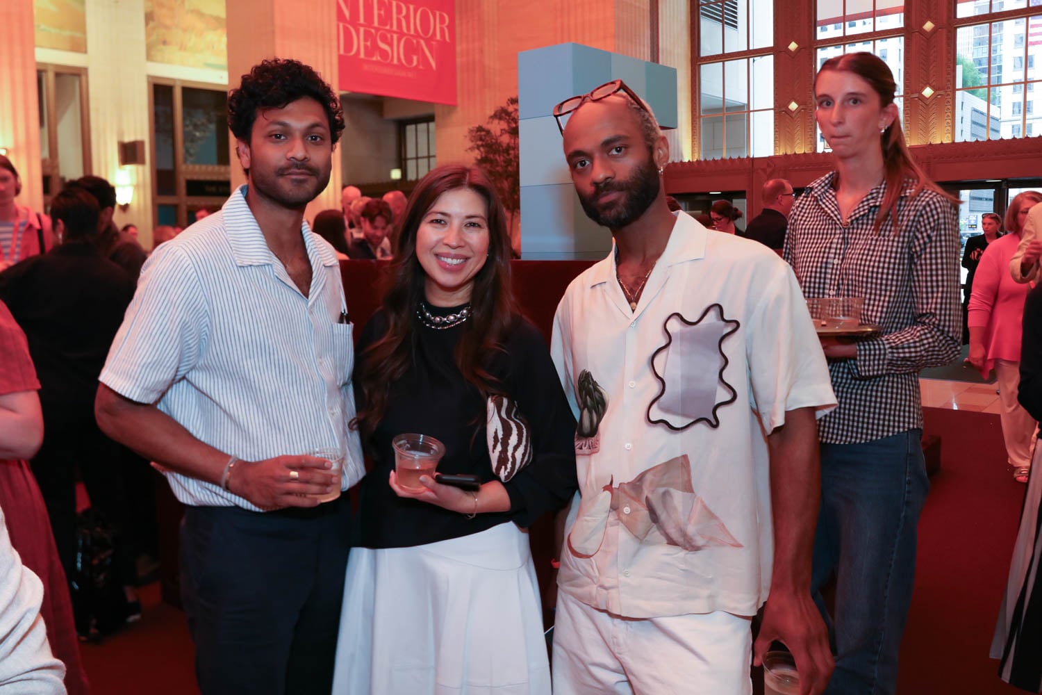 Three people stand together indoors at a social event, holding drinks and looking at the camera, with other attendees in the background.