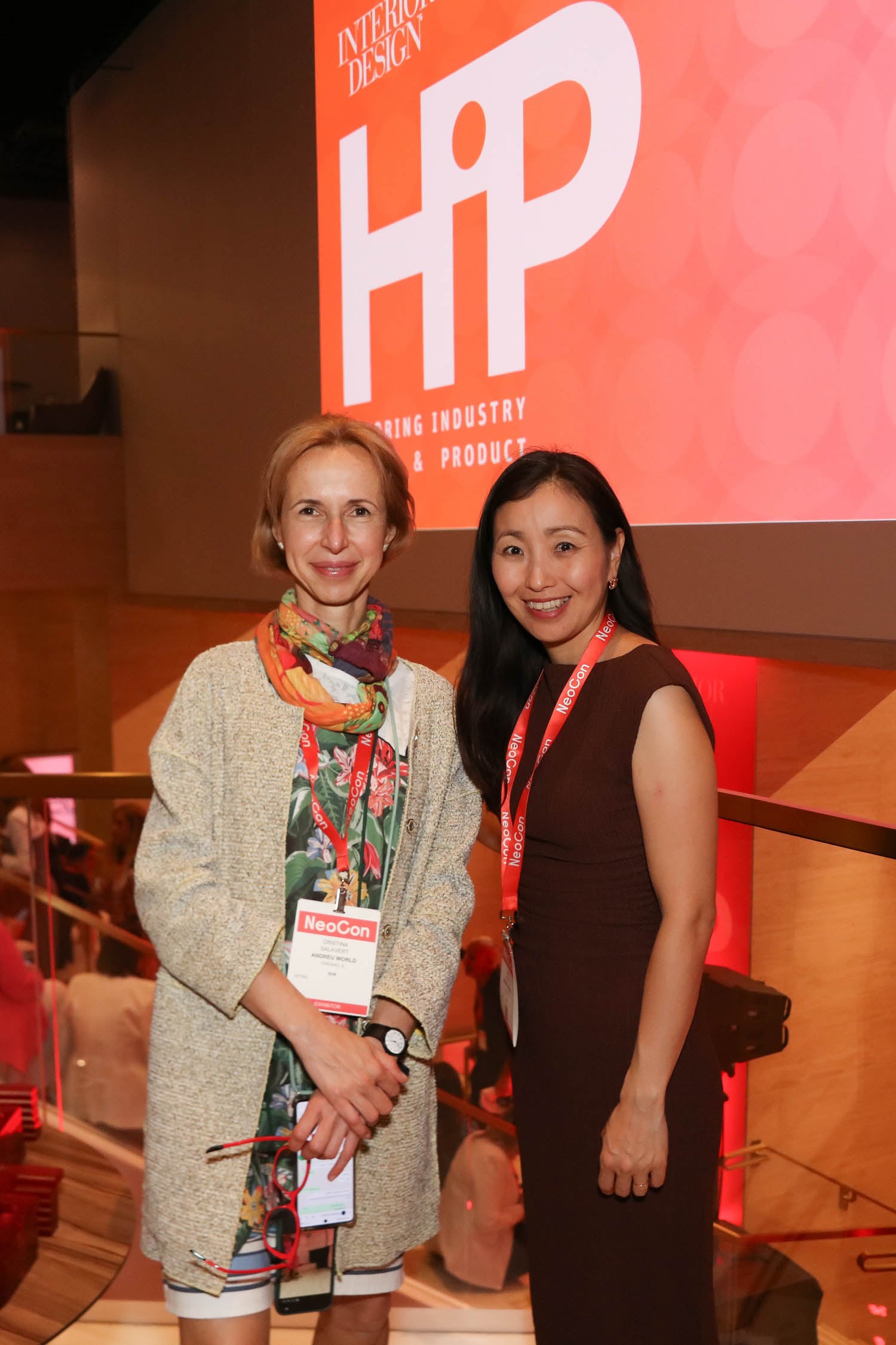 Two women pose and smile at the HiP Interior Design event, both wearing NeoCon badges, with a large event sign in the background.