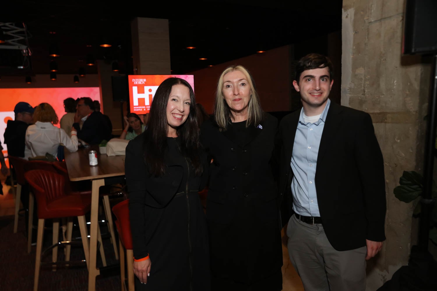 Three people pose and smile for a photo at an indoor event, with others socializing in the background near tables and a bright screen displaying text.