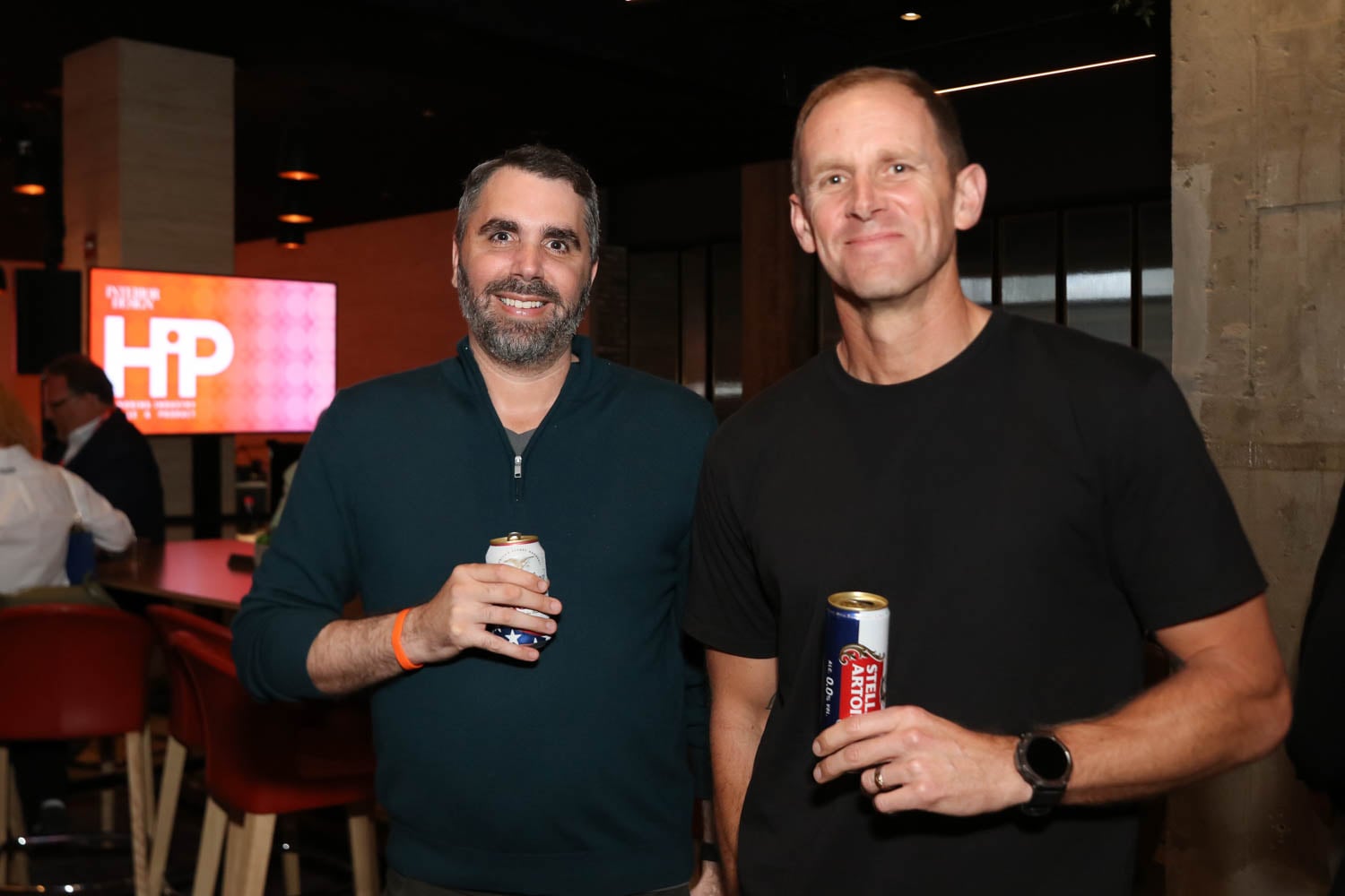 Two men stand indoors, each holding a beverage can, with a bar and a screen displaying "HiP" in the background.