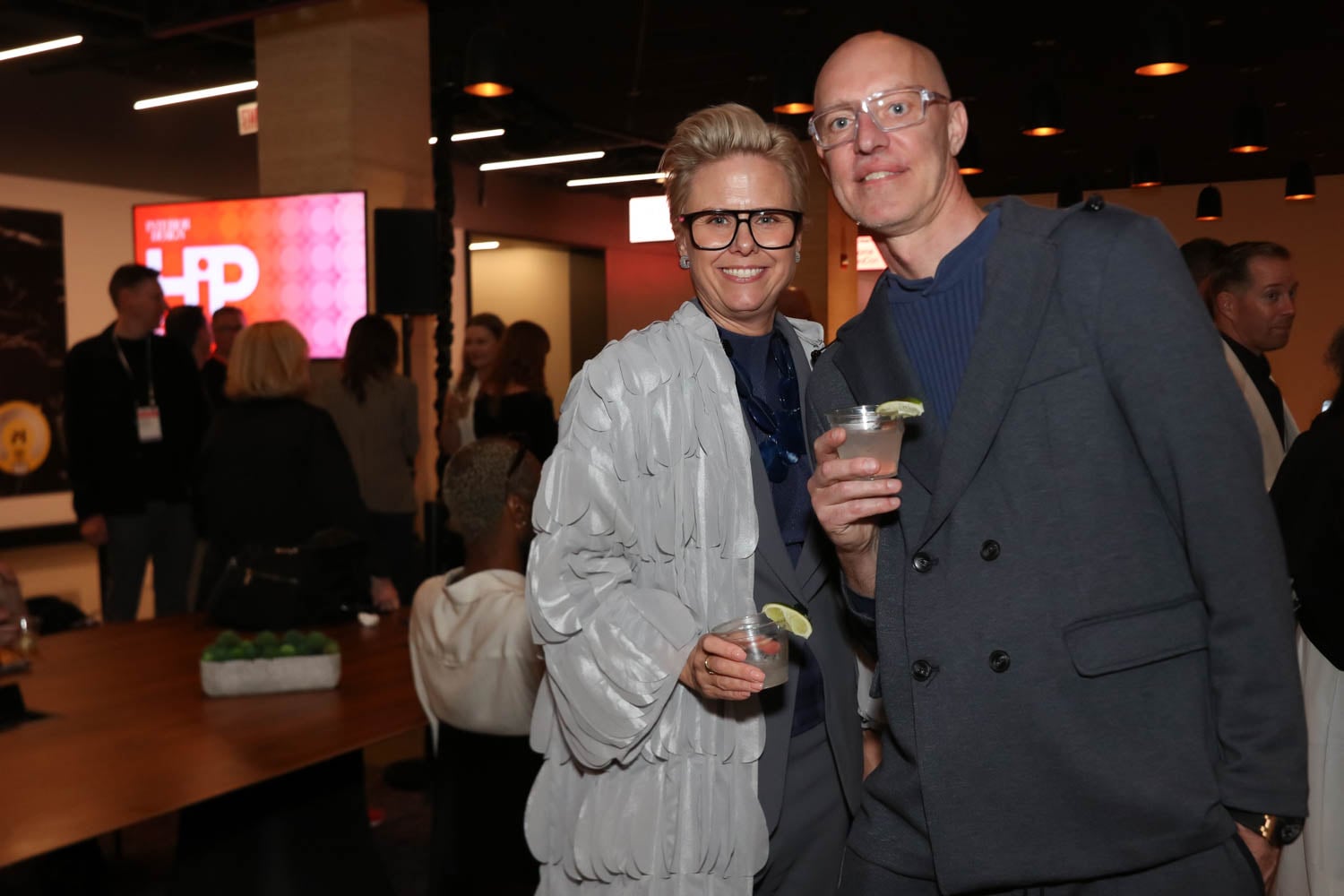 Two people stand close together holding drinks with lime wedges at a social event, with other attendees mingling in the background.