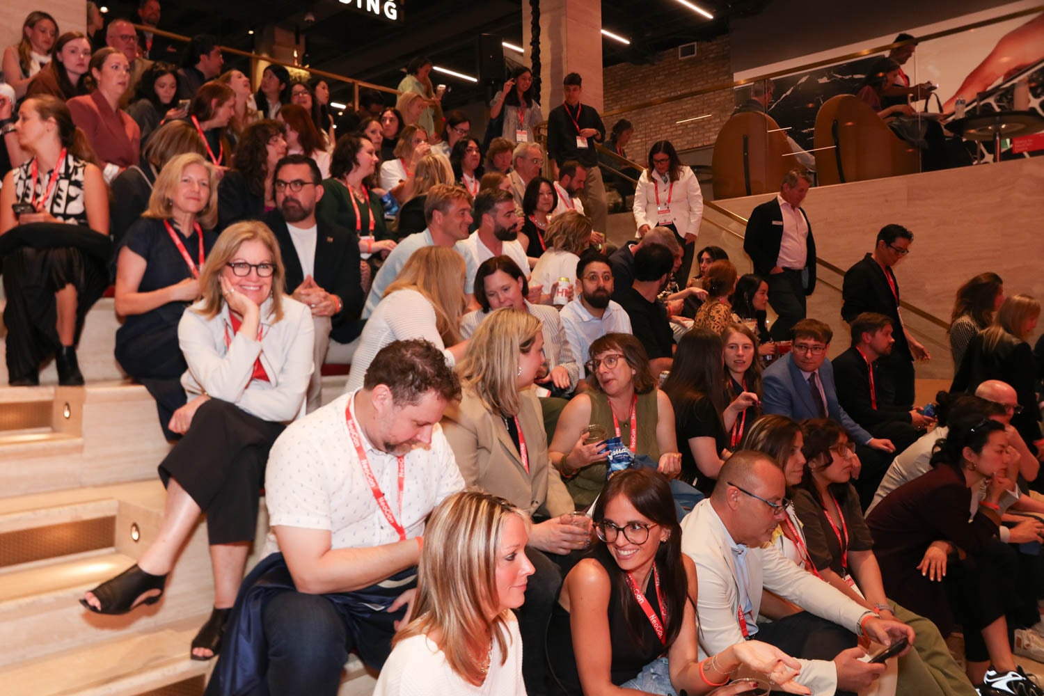 A large group of people wearing name badges are seated on tiered steps indoors, attending what appears to be a conference or event.