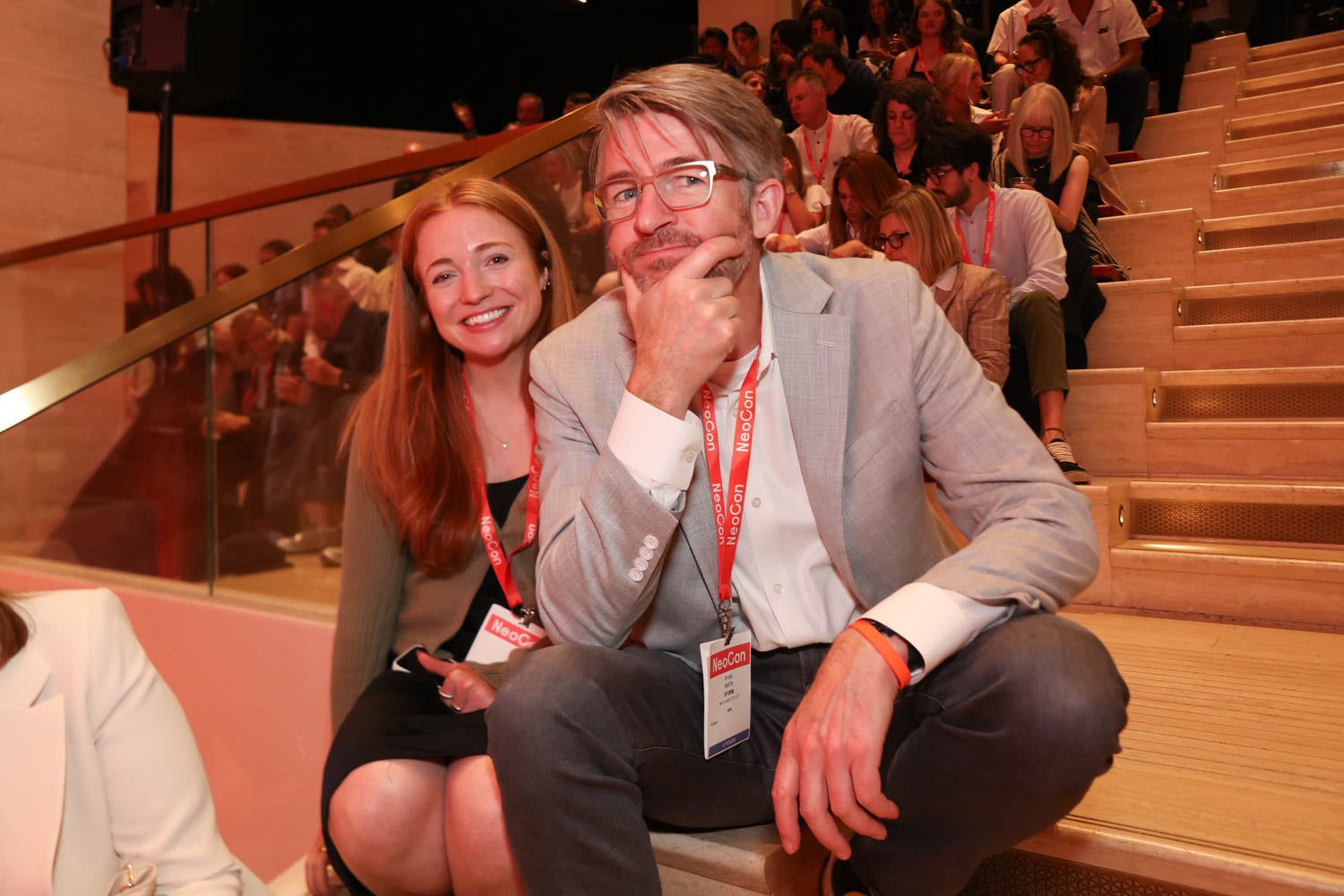 Two people with conference badges sit on steps among a crowd; the man in front is resting his chin on his hand, and the woman behind him is smiling at the camera.