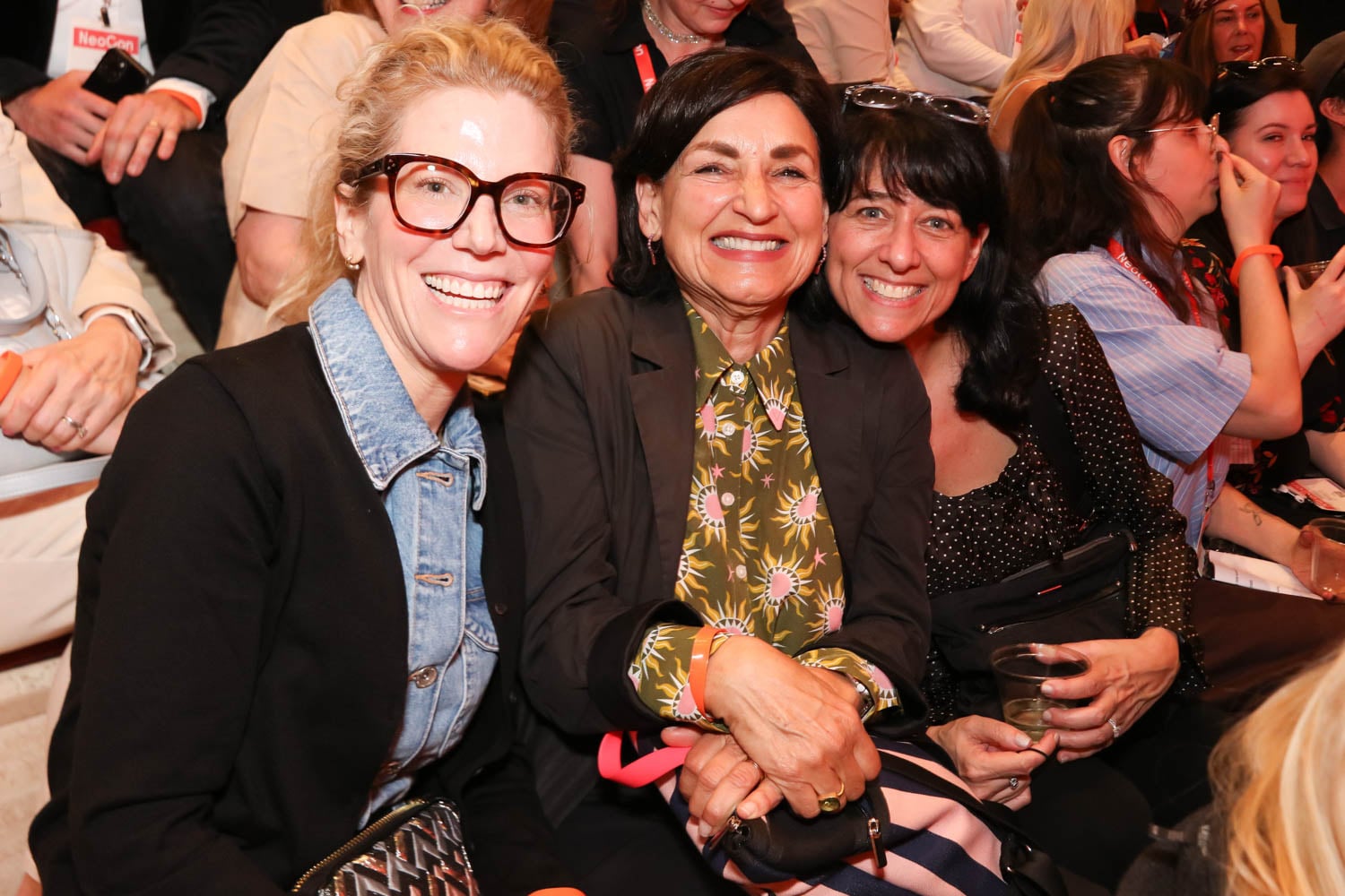 Three women sitting close together, smiling at the camera in a crowded indoor event with people seated around them.