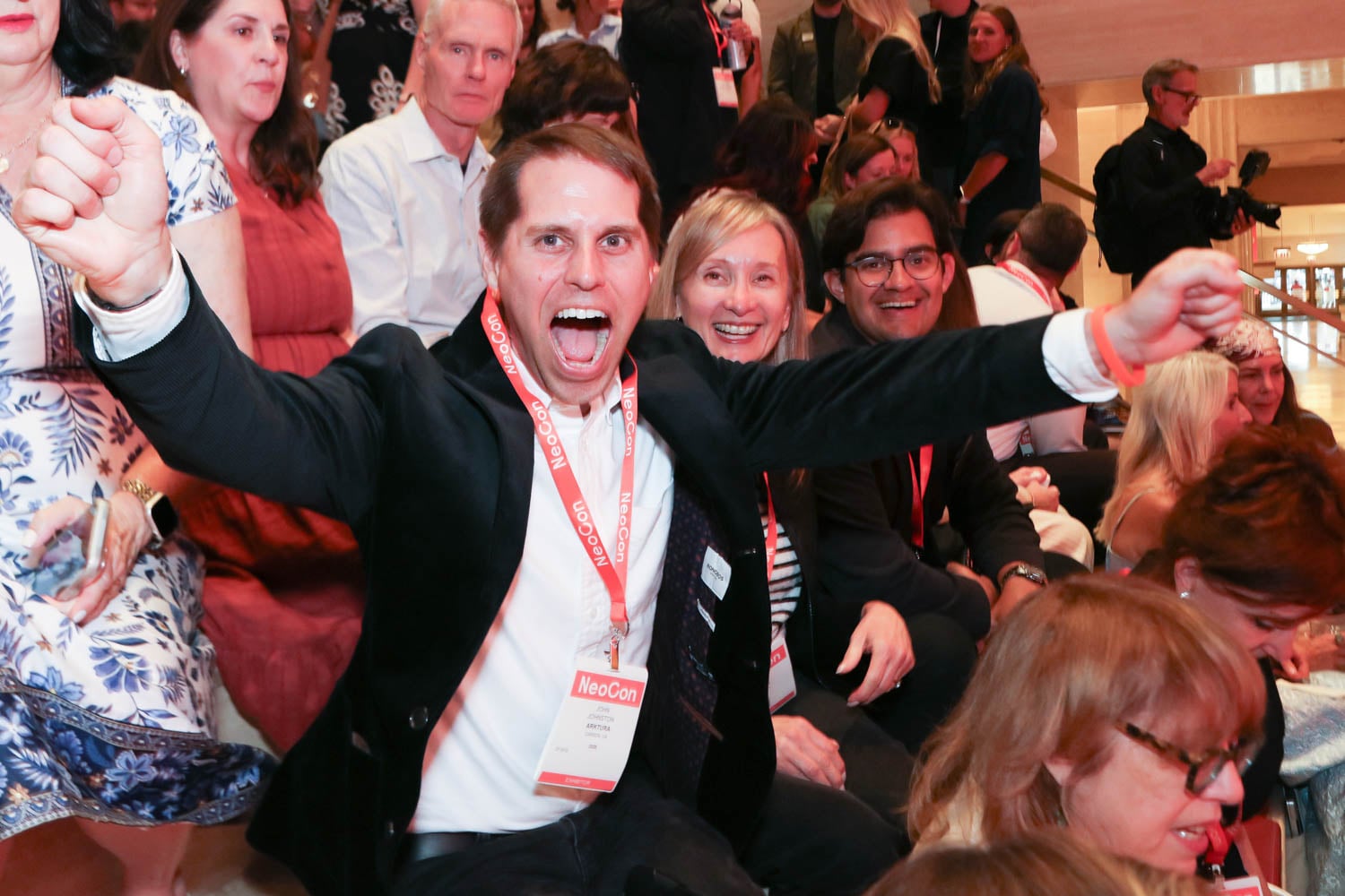 A group of people sit closely together at an indoor event; a man in front raises his arms and shouts excitedly, wearing a NeoCon badge around his neck.