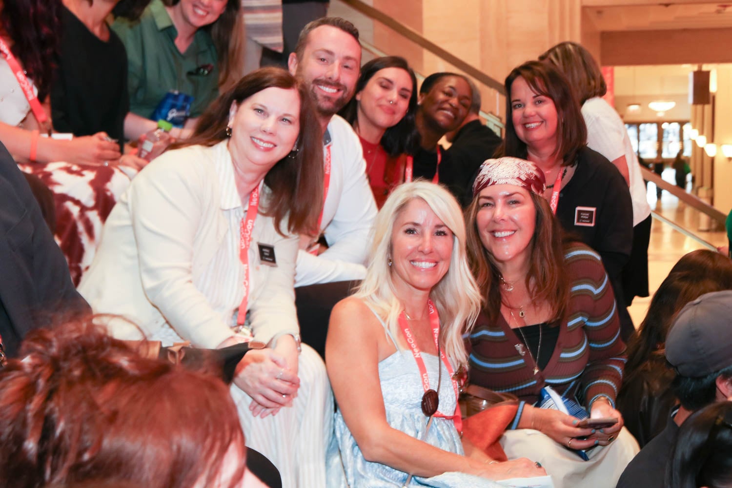 A group of people sit closely together on indoor steps, smiling at the camera and wearing badges on lanyards.