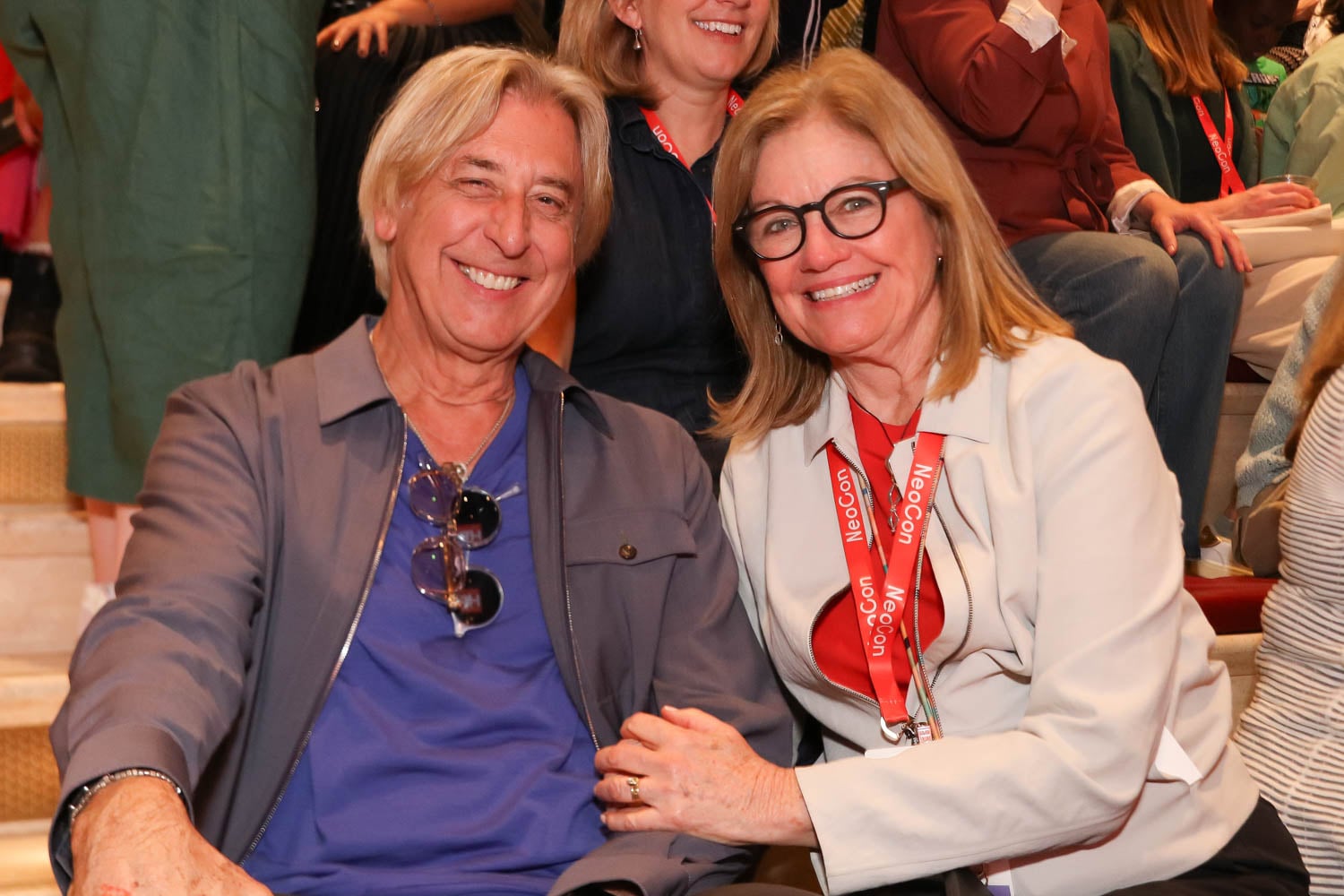 A man and a woman smiling and sitting close together in a crowd, both wearing event badges around their necks.
