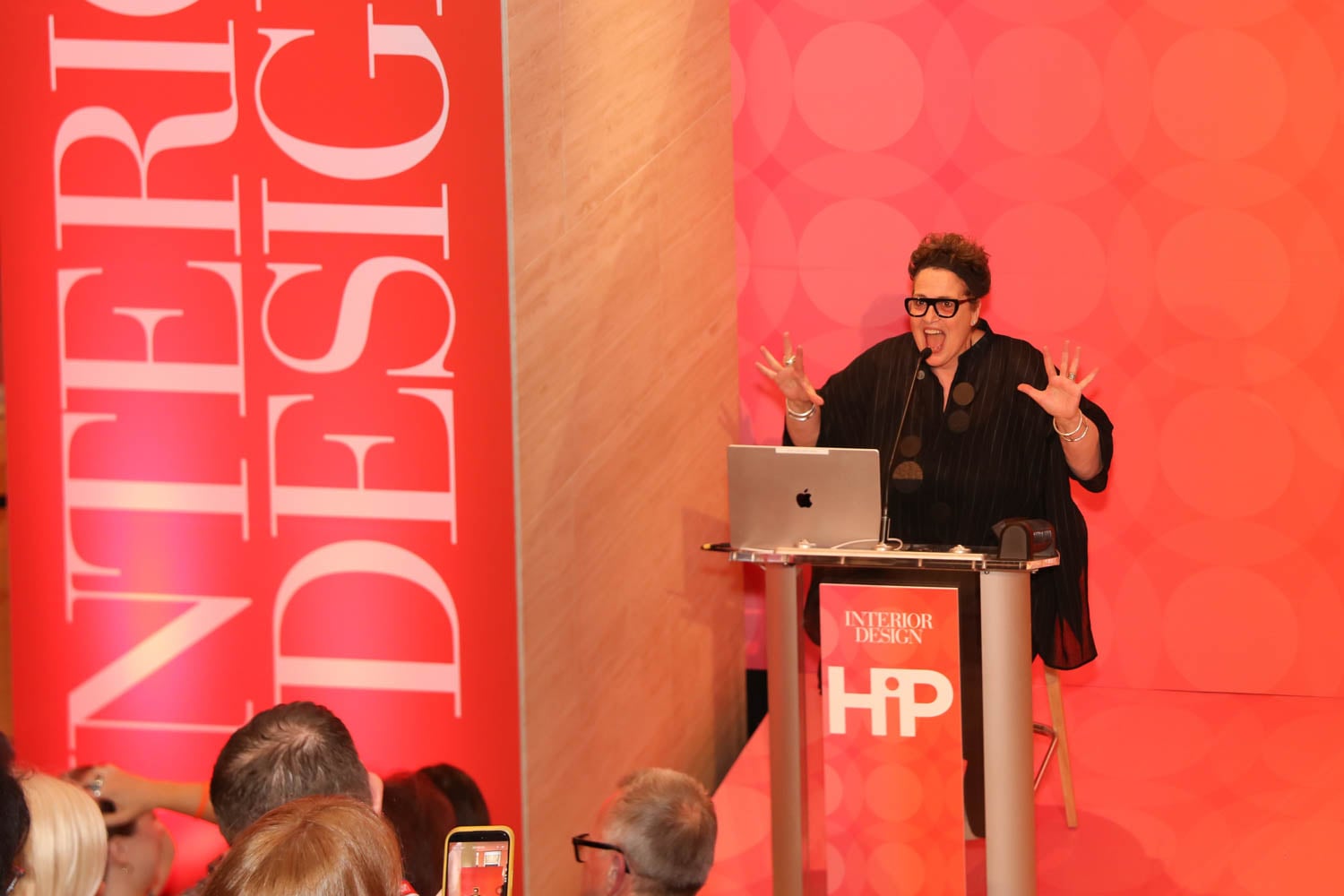 A person speaks enthusiastically at a podium labeled "HiP" and "Interior Design," with an audience watching in a brightly lit event space.