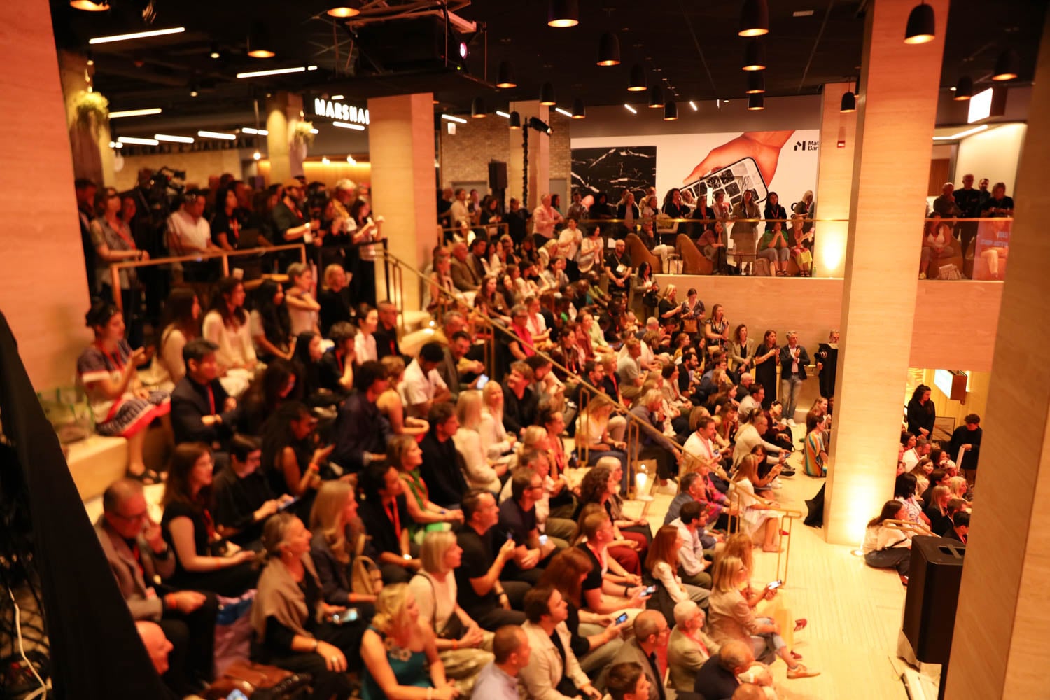 A large audience sits on tiered seating in a modern indoor venue, attentively watching an event or presentation.