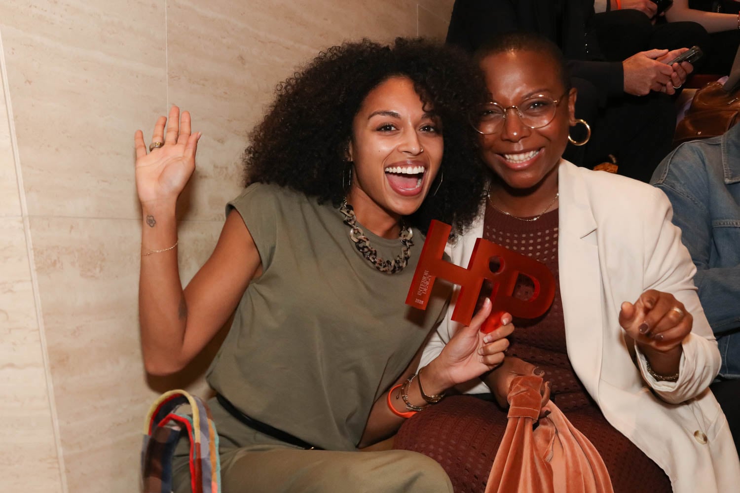Two women sitting together, smiling at the camera; one is holding a red "HPD" award and waving, both appear happy and casually dressed.