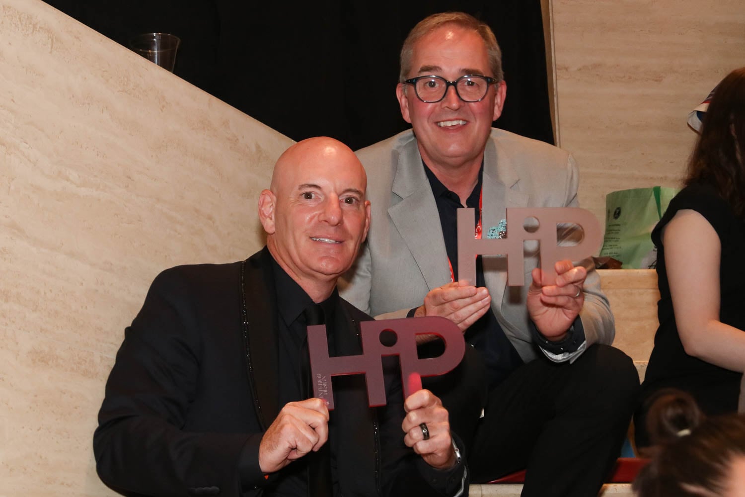 Two men in formal attire sit on stairs holding "HiP" award plaques and smiling at the camera.
